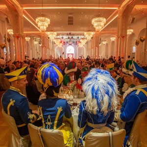 In der prunkvoll gestalteten Flora sitzen rund 500 Gäste an runden Tischen. Im Vordergrund sitzen Mitglieder der Tanzgruppen des Kölner Traditionskorps.