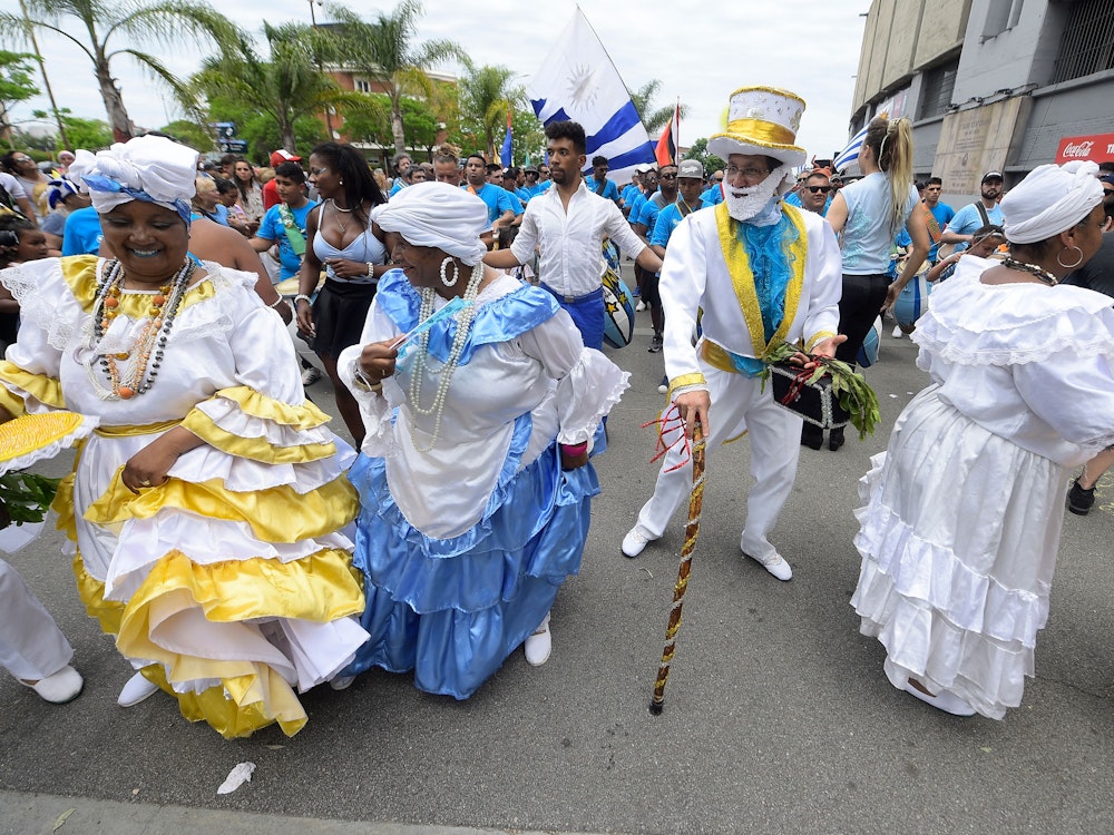 Fans aus Uruguay tanzen auf der Straße. Ein Mann trägt einen Anzug, die Frauen traditionelle Kleider – alle in den Landesfarben Gelb und Blau.