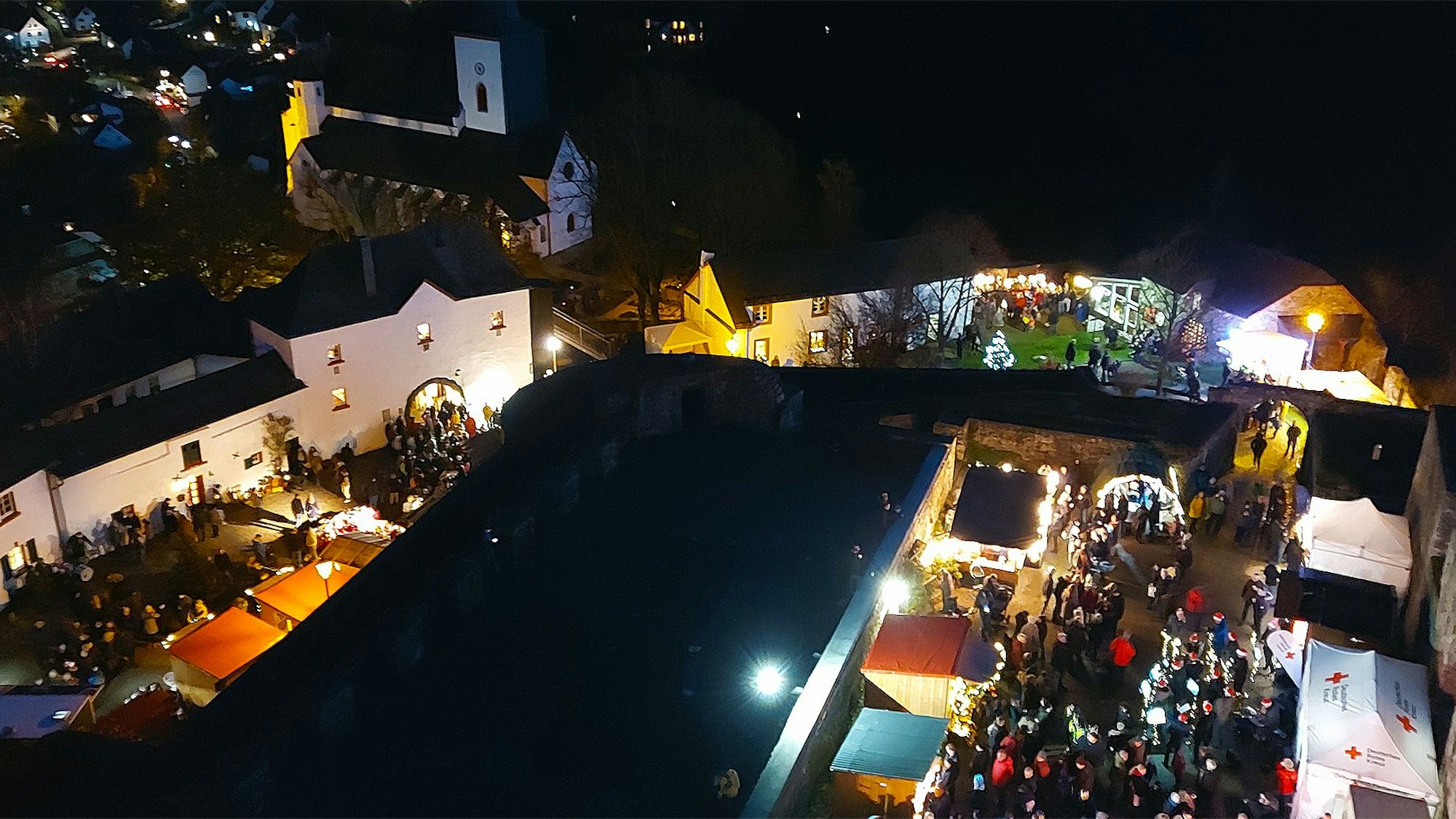 Vom Burgturm geht der Blick hinunter auf den Weihnachtsmarkt im mittelalterlichen Burgbering von Reifferscheid. In den illuminierten Gassen tummeln sich die Besucher zwischen den Ständen, im Hintergrund sieht man die Pfarrkirche und im Tal die Häuser von Reifferscheid.