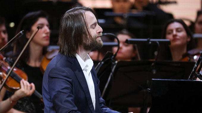Der Pianist trägt lange Haare und Bart. Er spielt auf einem Steinway Flügel, im Hintergrund sind weitere Musiker zu sehen.