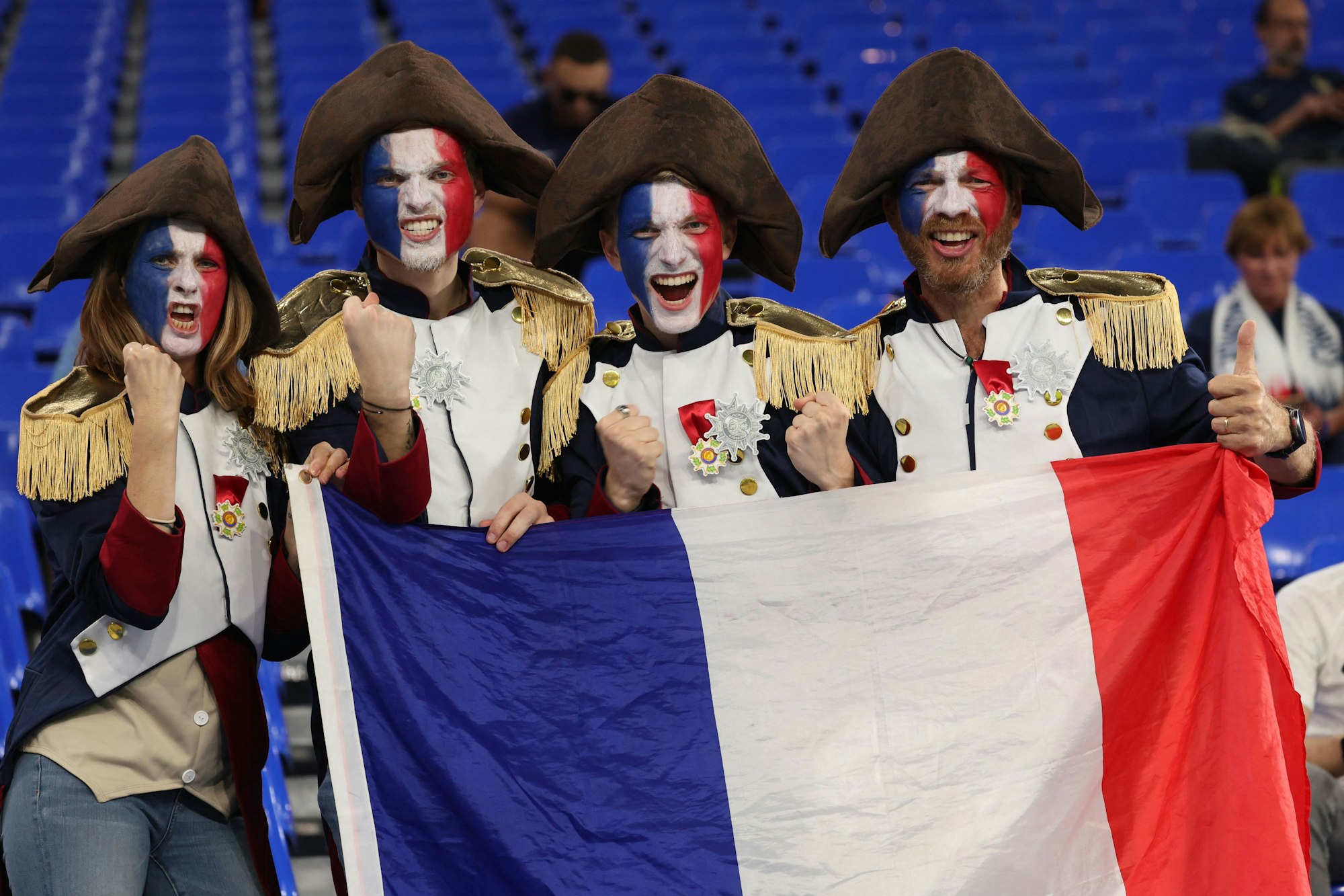 Vier Frankreich-Fans posieren in ihren Kostümen und einer Frankreich-Flagge auf der Tribüne.