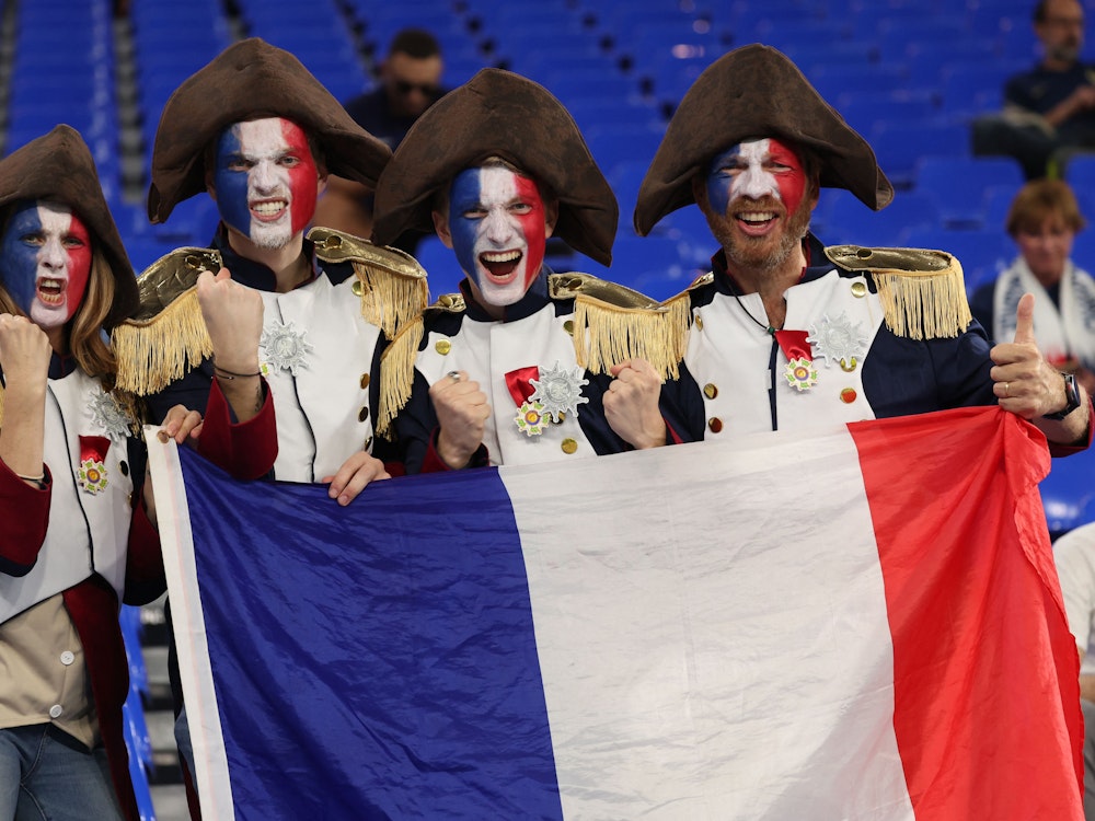 Vier Frankreich-Fans posieren in ihren Kostümen und einer Frankreich-Flagge auf der Tribüne.
