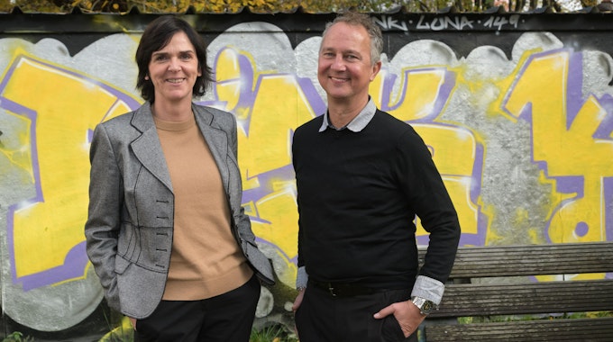 Sigrid Schule und André Eigenbrod, die neuen Leiter der Kreaschule in Bergisch Gladbach, stehen vor einer mit gelb-lilanen Graffitis verzierten Wand.