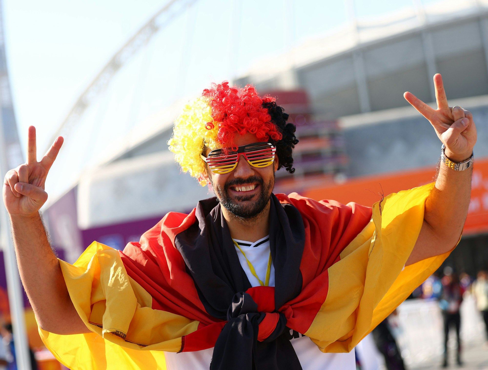 Ein Deutschland-Fan mit Flagge und Perücke steht vor einem Stadion in Katar und zeigt mit beiden Händen ein Peace-Zeichen.