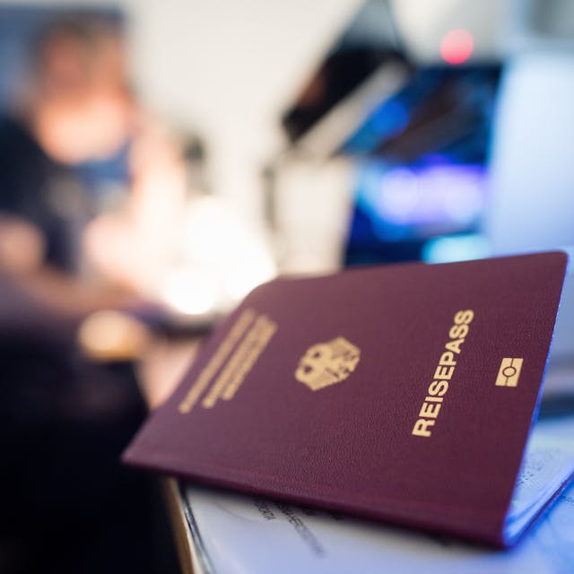 Ein deutscher Reisepass liegt auf einem Tisch, blaues Licht scheint darauf, eine Person ist verschwommen im Hintergrund zu sehen.