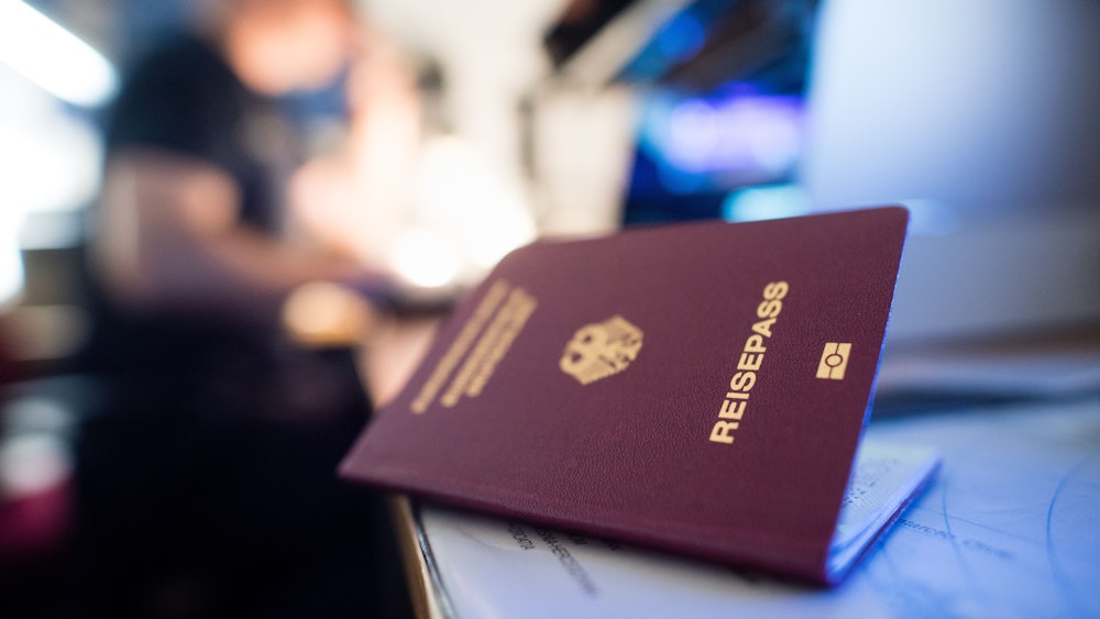 Ein deutscher Reisepass liegt auf einem Tisch, blaues Licht scheint darauf, eine Person ist verschwommen im Hintergrund zu sehen.