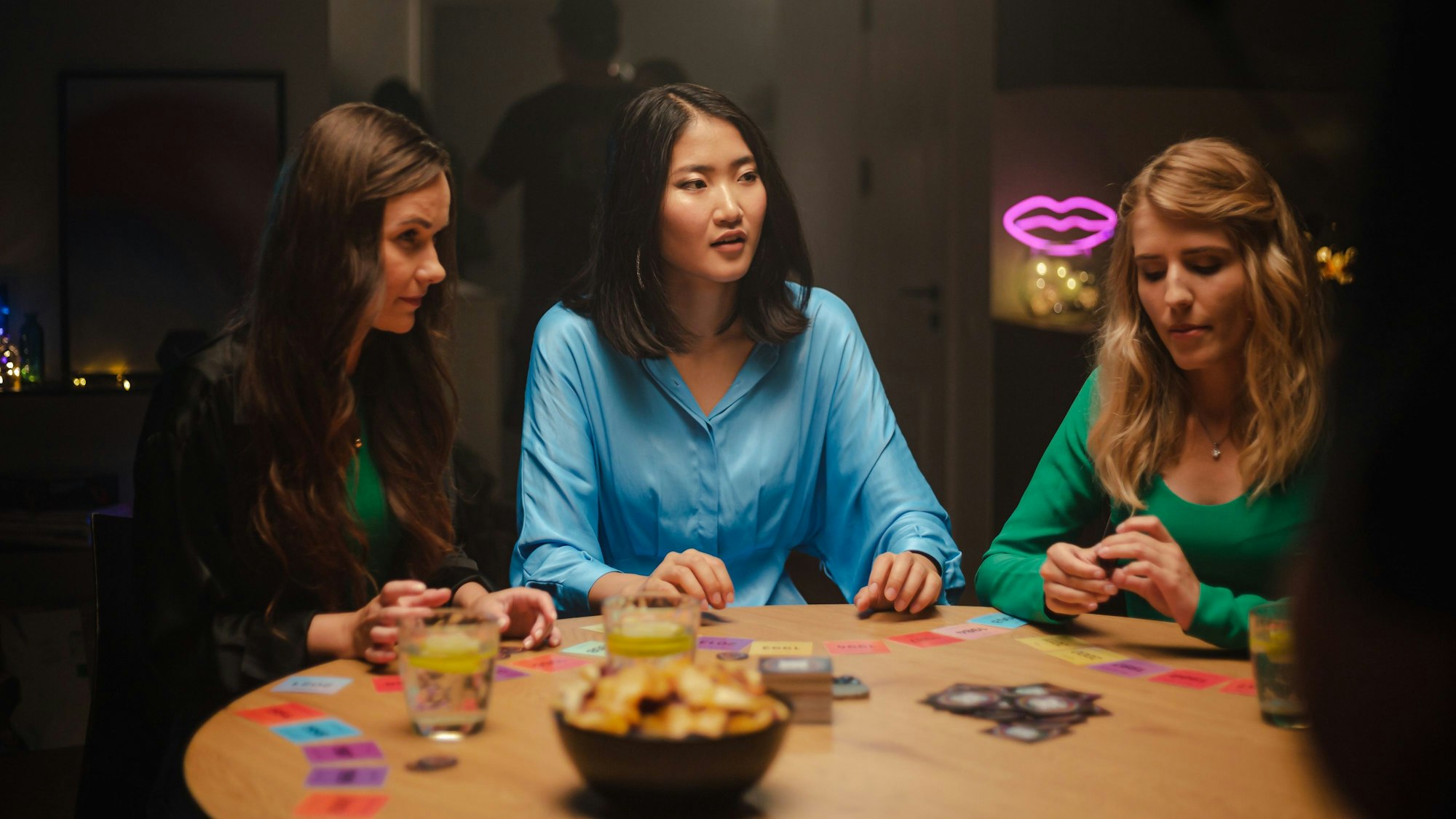 drei Frauen sitzen an einem runden Holztisch, vor ihnen liegen Spielkarten