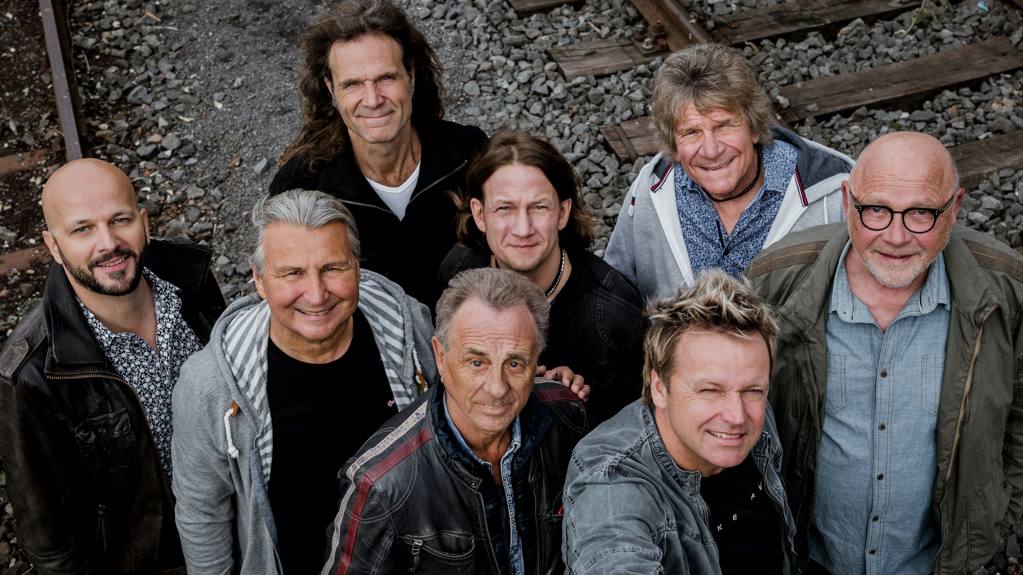 Die acht Bandmitglieder stehen auf einer Bahntrasse, von oben aufgenommen.