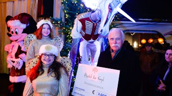 „wir helfen“-Geschäftsführer Karl-Heinz Goßmann sthet mit einem symbolischen Scheck in Höhe von 3000 Euro auf der Bühne des Markts der Engel umgeben von Engeln, einer Mikey Maus und einem Einhorn auf Stelzen.