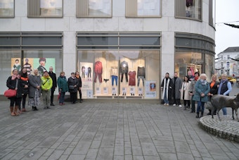 Rechts und links eines Schaufensters, in dem Kleidungsstücke missbrauchter Frauen hängen, stehen die Beteiligten der Ausstellung.