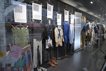 In einem Kleidungsgeschäft in Bergisch Gladbach sind an einer Wand verschiedene Kleidungsstücke ausgestellt. Über ihnen hängt jeweils ein großes Blatt Papier mit QR-Code.