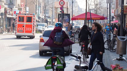 Unübersichtlicher Straßenverkehr auf der Venloer Straße. Ein Krankenwagen fährt auf der Straße, eine Fahrradfahrerin und eine Fußgängerin kreuzen sich.