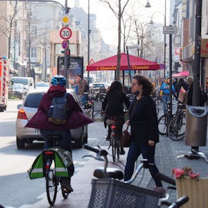 Unübersichtlicher Straßenverkehr auf der Venloer Straße. Ein Krankenwagen fährt auf der Straße, eine Fahrradfahrerin und eine Fußgängerin kreuzen sich.