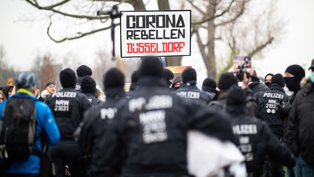 Demonstranten halten bei einer Demonstration am Rheinufer in Düsseldorf ein Schild mit der Aufschrift „Corona Rebellen Düsseldorf“ in die Höhe, davor stehen Einsatzkräfte der Polizei.