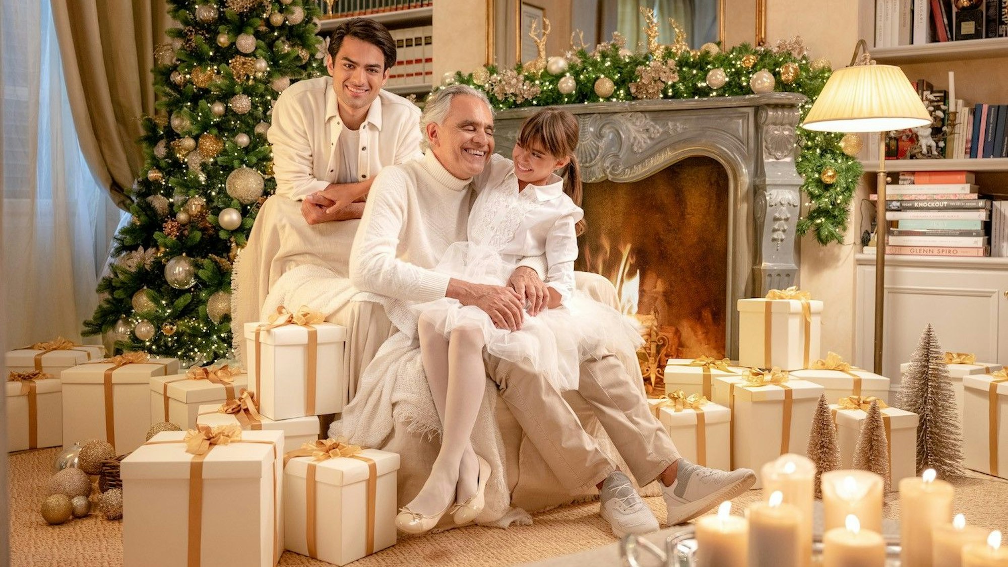 Andrea Bocelli an Weihnachten mit Familie, links ein großer, reichlich geschmückter Weihnachtsbaum, davor Weißgold verpackte Geschenke. Die Kerzen leuchten und alle strahlen.