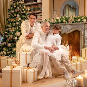 Andrea Bocelli an Weihnachten mit Familie, links ein großer, reichlich geschmückter Weihnachtsbaum, davor Weißgold verpackte Geschenke. Die Kerzen leuchten und alle strahlen.