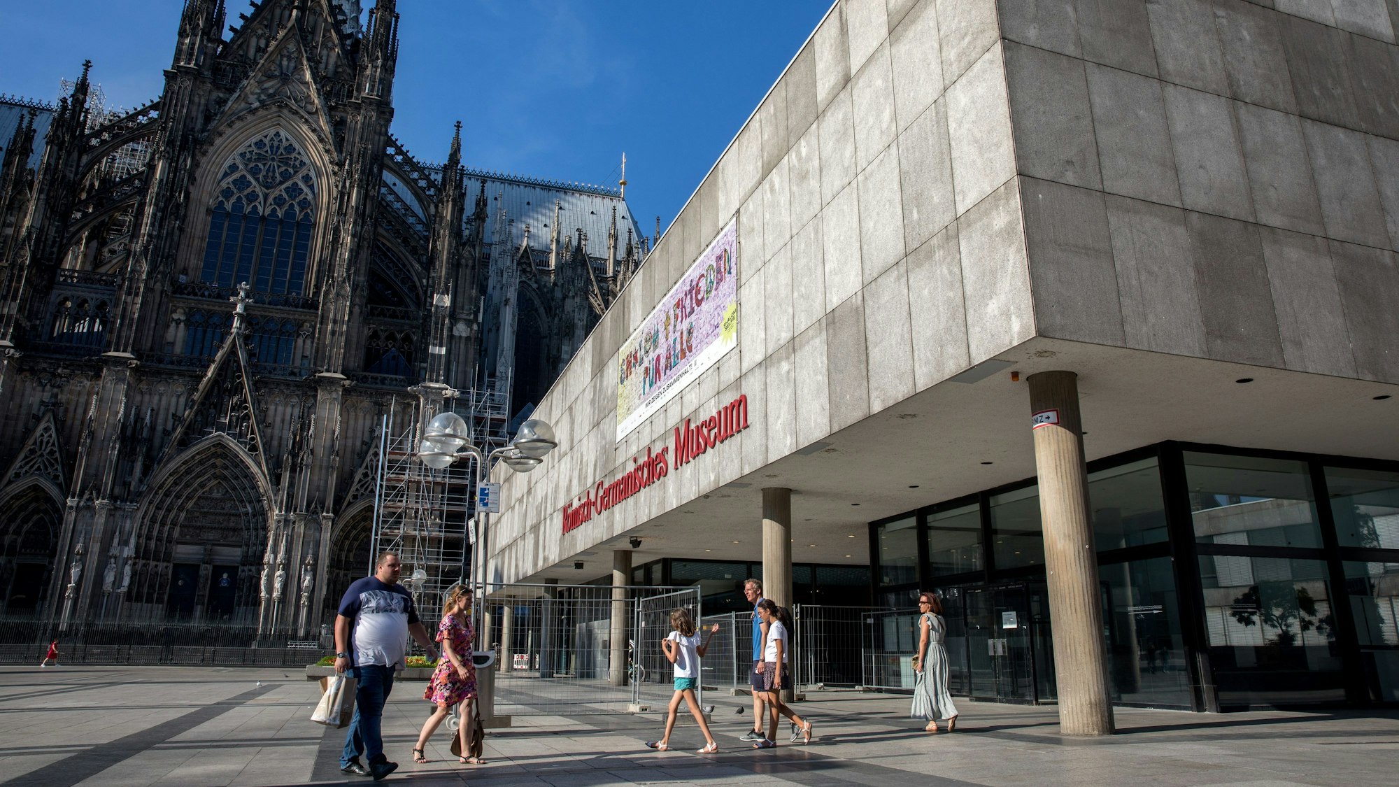 Menschen laufen über die Domplatte am Dom und dem Römisch-Germanischen Museum vorbei