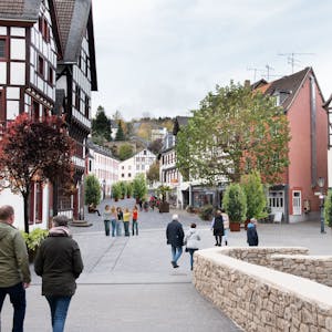 Das Bild zeigt den Marktplatz von Bad Münstereifel mit der Marktstraße im Hintergrund mit Blick von der Jesuitenkirche über die Heinz-Küpper-Brücke aus. Der Fotografie wurden Pflanzkübel und Bänke auf der Marktstraße hinzugefügt, die Autos wurden entfernt.