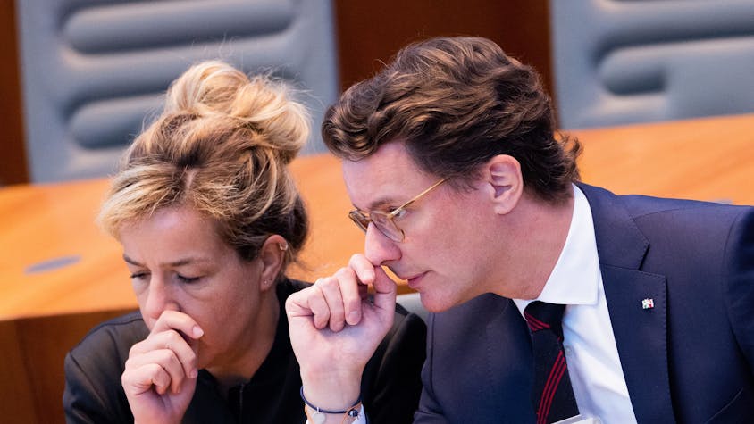 Die NRW-Politiker Mona Neubaur und Hendrik Wüst sitzen im Landtag, sie gucken besorgt, beide halten sich die Hände vor das Gesicht.