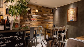 Die Inneneinrichtung des Café 1980, eine bunte Holzwand und einige Tische mit schlichten Stühlen sind zu sehen.