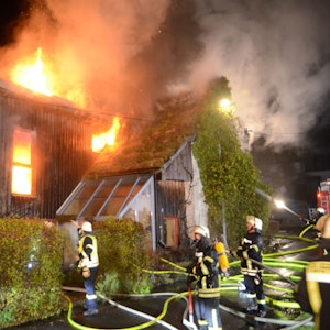 Feuerwehrleute stehen vor einem Haus mit Holzverkleidung, das in Flammen steht. Das Feuer kommt aus einem Fenster und dem Dach. In einem anderen Fenster ist deutlich zusehen, wie es im Inneren des Hauses brennt.