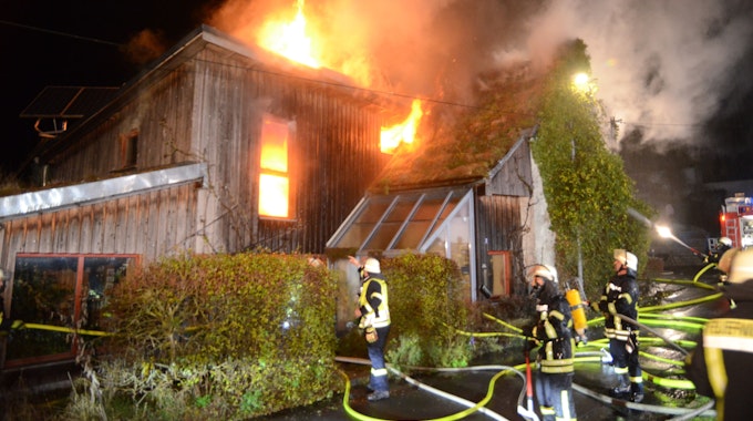 Feuerwehrleute stehen vor einem Haus mit Holzverkleidung, das in Flammen steht. Das Feuer kommt aus einem Fenster und dem Dach. In einem anderen Fenster ist deutlich zusehen, wie es im Inneren des Hauses brennt.