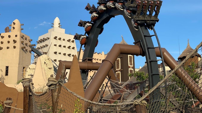 Eine Achterbahn hängt in einem Looping über Kopf. Im Hintergrund stehen Gebäude aus dem Themenreich Afrika im Phantasialand.