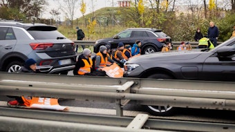 In der Bildmitte sitzen Klimaaktivisten in orangen Leuchtwesten auf der gesamten Fahrbahnbreite einer Autobahn in einer Reihe und blockieren so den Verkehr. Vor und hinter ihnen stehen Autos.