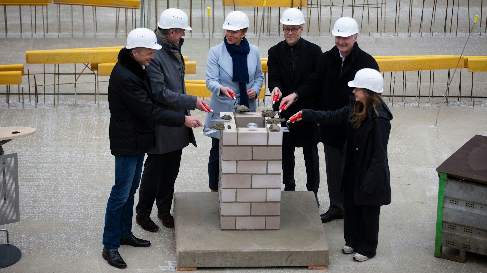 Sechs Personen stehen mit Maurerkellen mit Zement an dem Grundstein, auf den gleich die Grundsteinplatte gelegt wird.