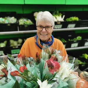 Ingrid Bauer steht vor einem Regal mit Gemüse. Sie hält einen Blumenstrauß in der Hand.