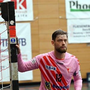 Ein Handballtorwart blickt aufs Spielfeld und hält sich mit dem rechten Arm am Pfosten fest.