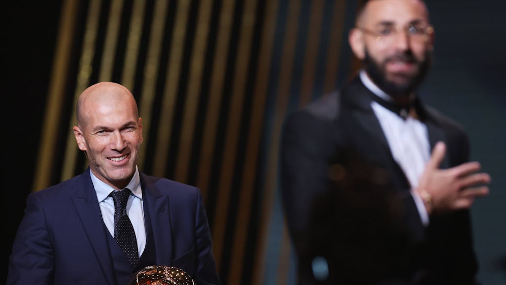 Zinedine Zidane hält die Trophäe von Ballon-d'or-Sieger Karim Benzema in der Hand und lächelt.