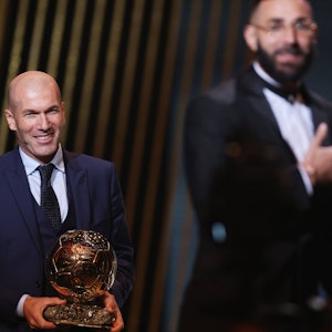 Zinedine Zidane hält die Trophäe von Ballon-d'or-Sieger Karim Benzema in der Hand und lächelt.