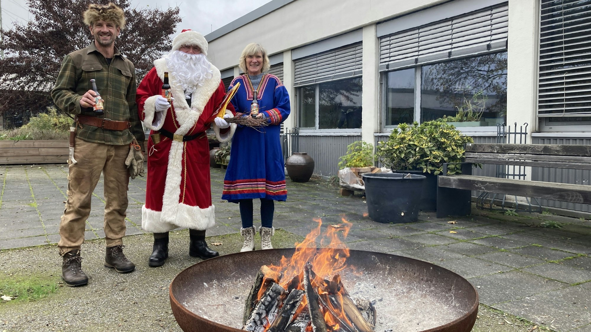 Trapper, Weihnachtsmann und Samifrau stehen in Verkleidung vor einer großen Feuerschale.