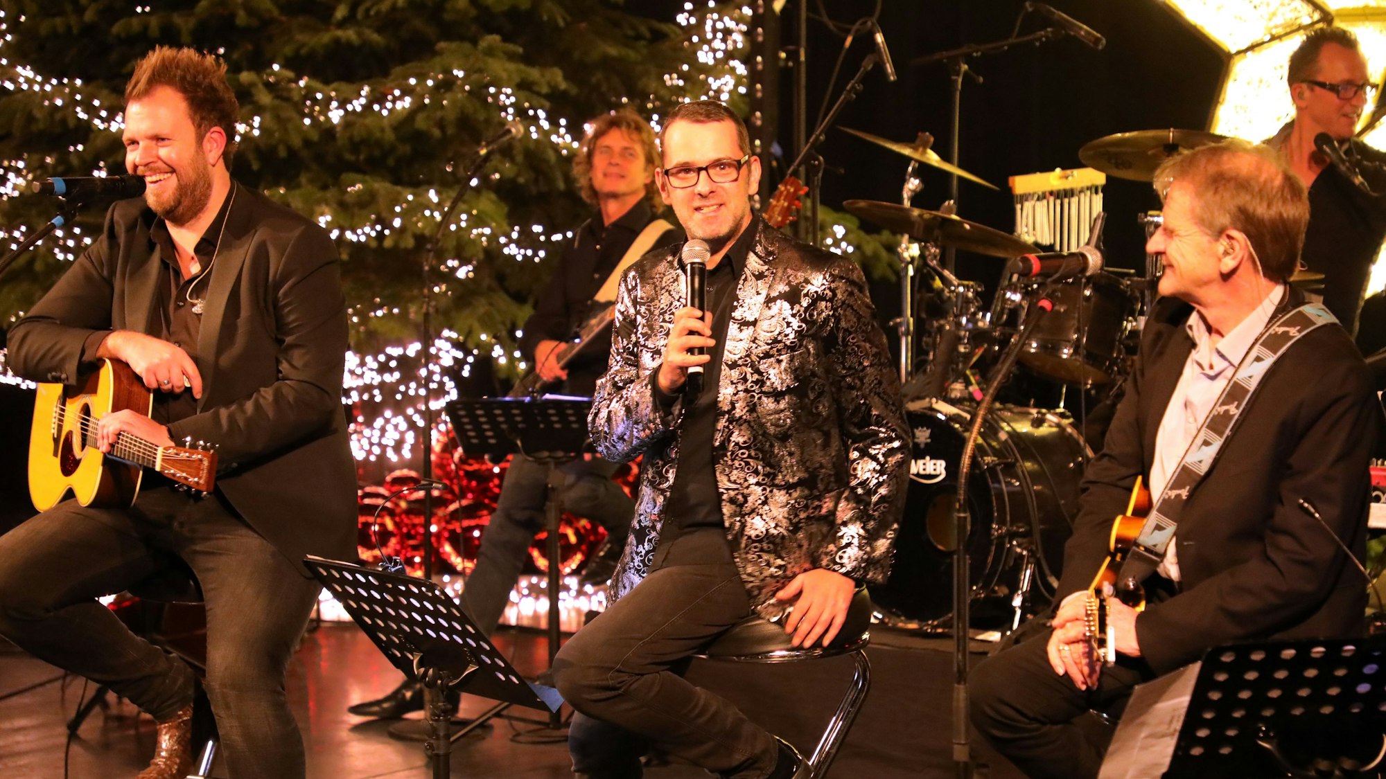Der Komödiant Marc Metzger sitzt zwischen Musikern der Paveier auf einer weihnachtlich geschmückten Bühne.