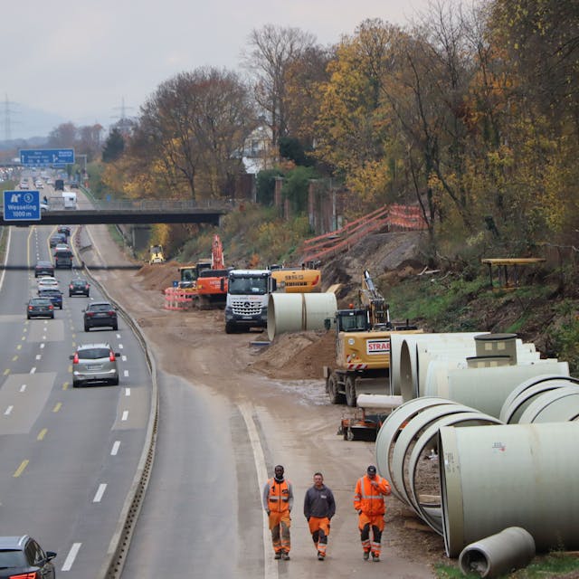 Autos fahren auf der Autobahn 555 an einer Baustelle vorbei. Am rechten Fahrbahnrand laufen Bauarbeiter an Kanal-Bauteilen vorbei.
