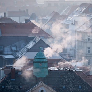 Die Dächer einer Siedlung im Winter, aus den Schornsteinen strömt Gas.