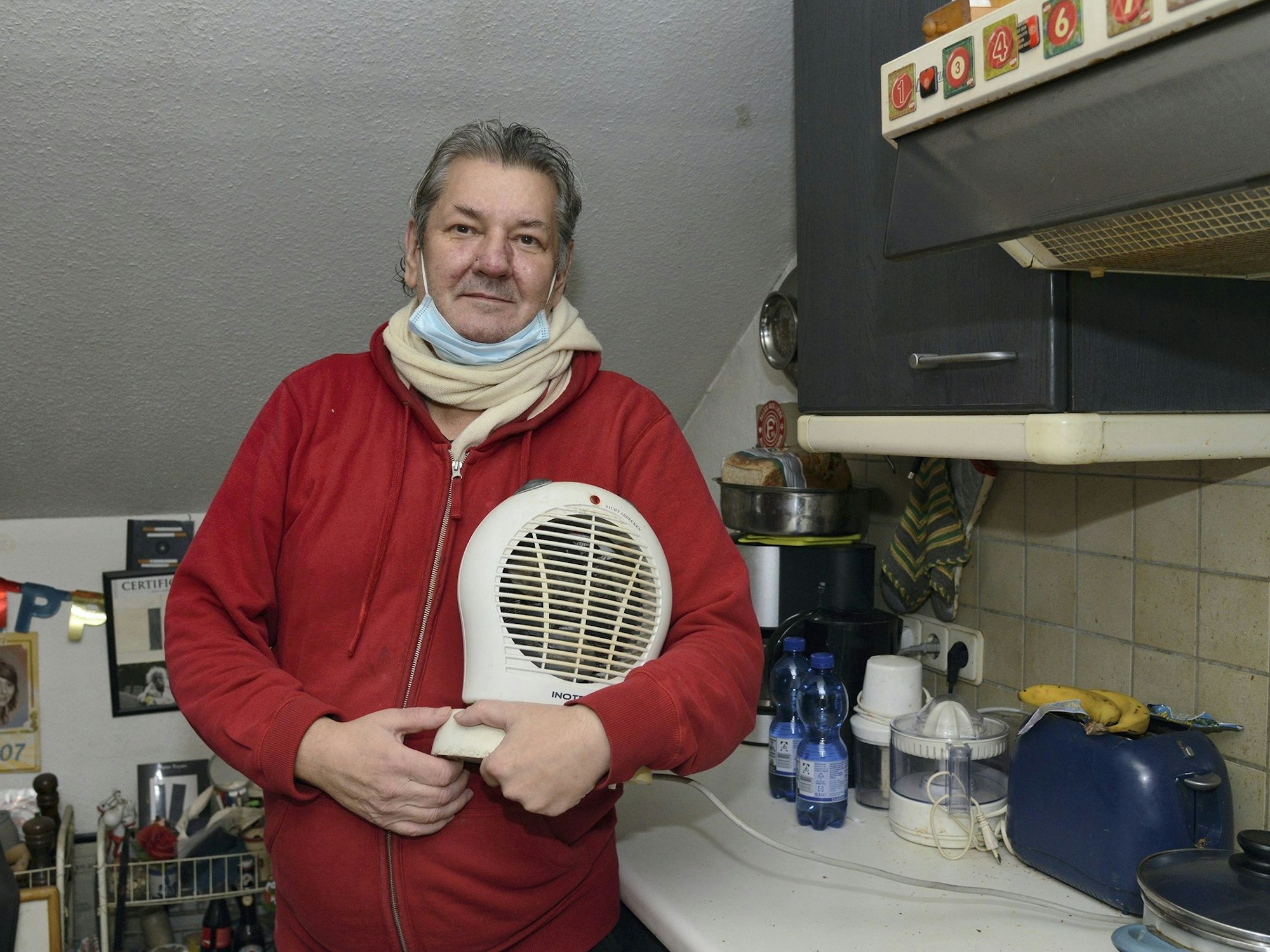 Harald Berndt steht mit Schal in seiner Küche und hält einen Heizlüfter in den Händen.