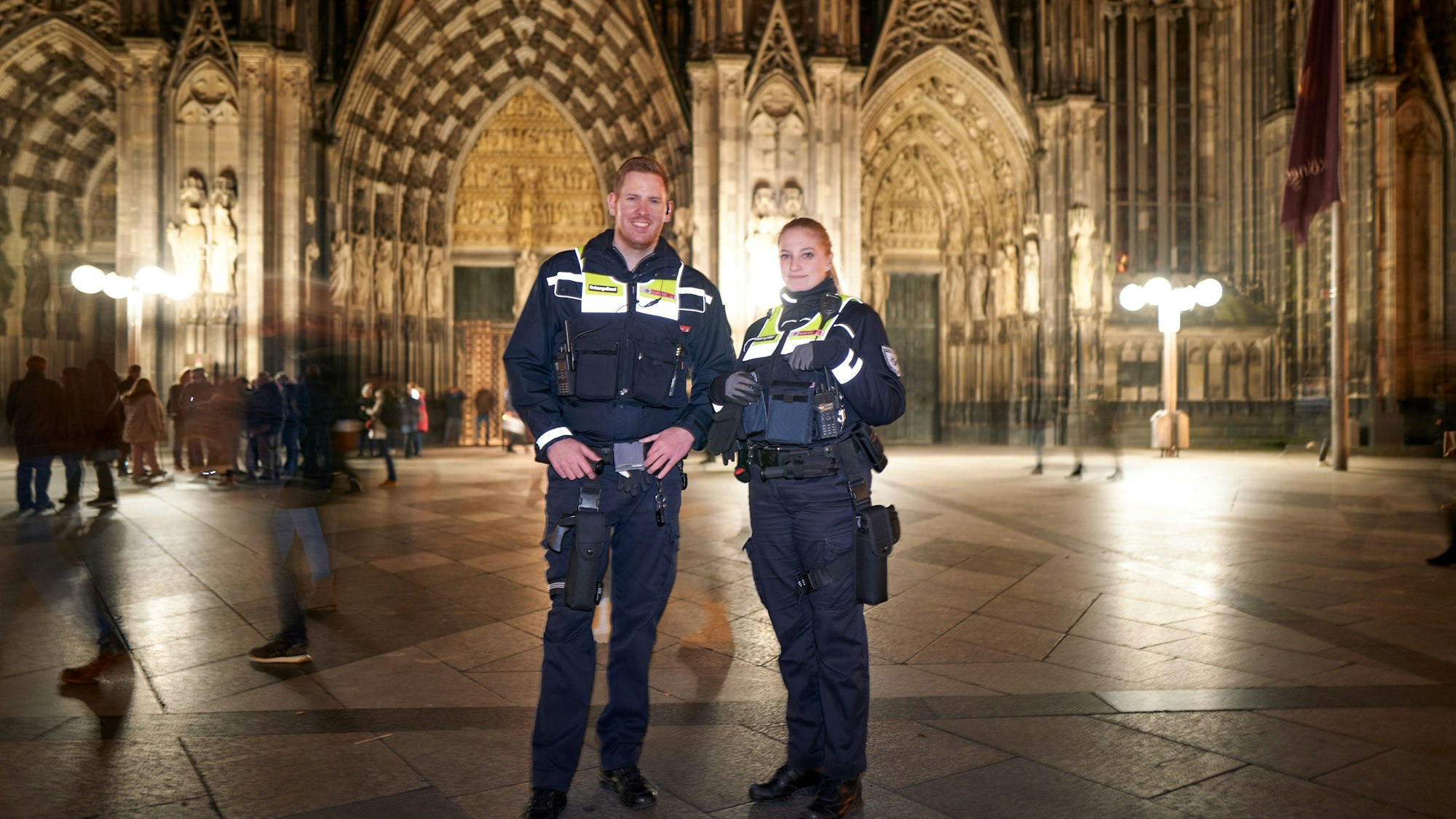 Die beiden Mitarbeitenden des Kölner Ordnungsamtes am Abend auf der Domplatte. Im Hintergrund steht der hell erleuchtete Kölner Dom.