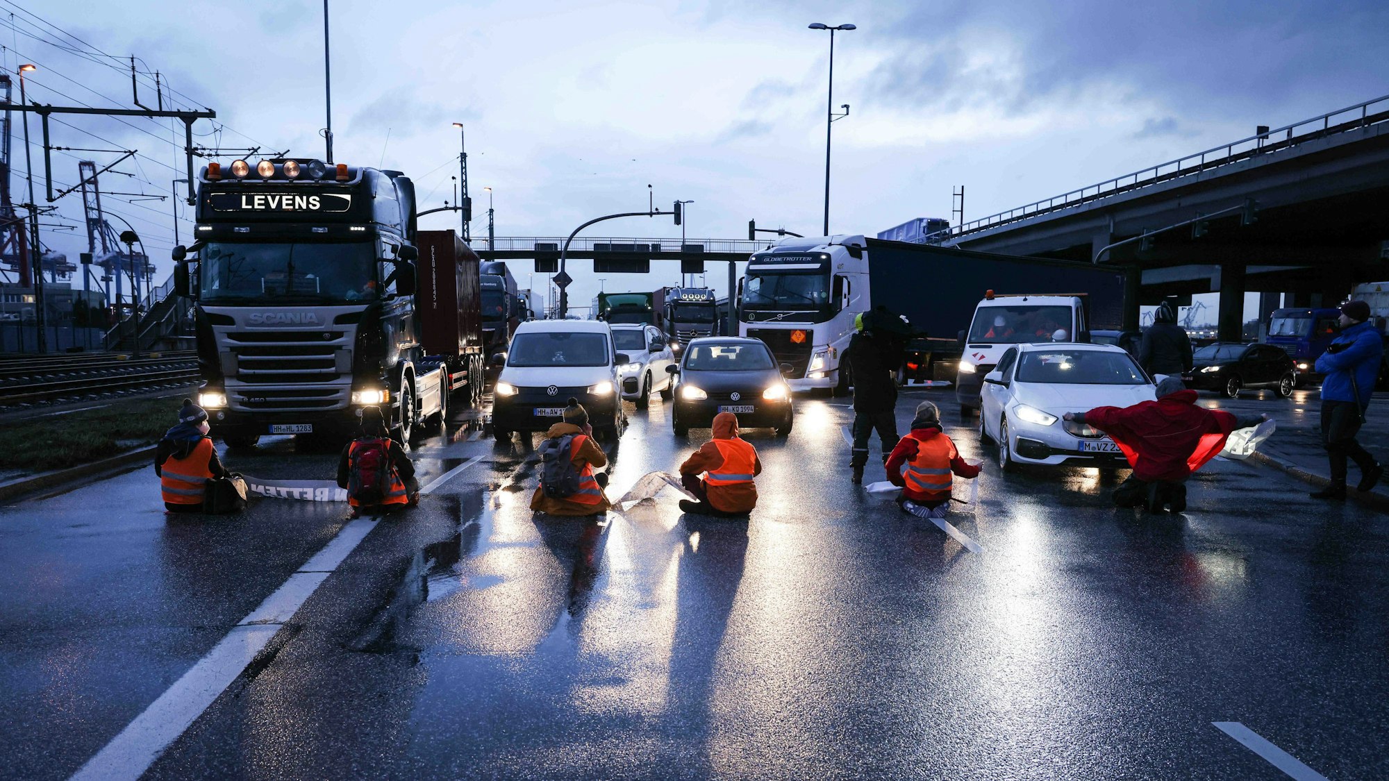 Klimaaktivisten der Aktion „Aufstand der letzten Generation“ blockieren am Morgen eine Kreuzung am Zollamt Waltershof zur Köhlbrandbrücke und zur Autobahn A7. Sie tragen Warnwesten und sitzen auf der Straße. Autos stehen vor ihnen.