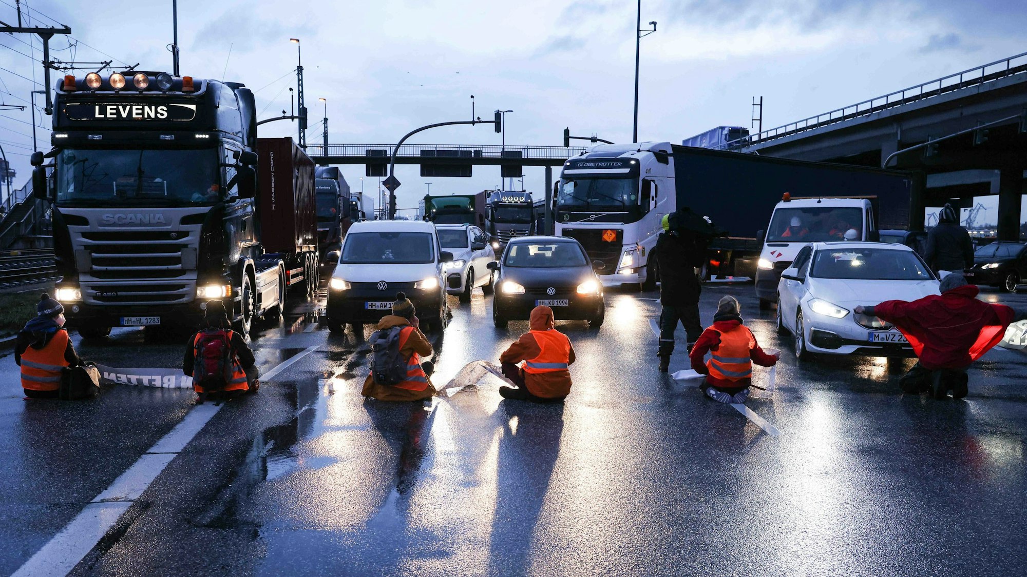 Klimaaktivisten sitzen mit dem Rücken zur Kamera auf einer mehrspurigen Kreuzung. Vor ihnen stehen Autos und LKW.
