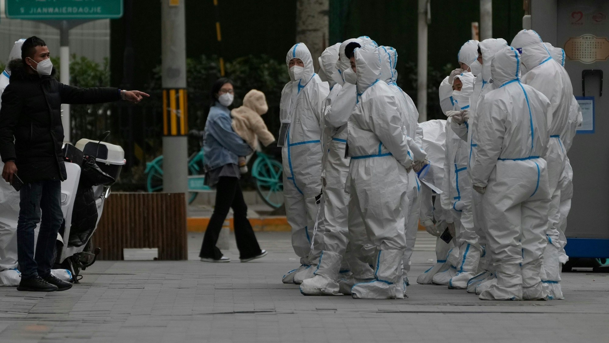 China: Arbeiter in Corona-Schutzkleidung versammeln sich zu ihrem Dienst. Sie stehen als Gruppe auf der rechten Seite des Bildes. Auf der linken Seite steht ein Mann in ziviler Kleidung.