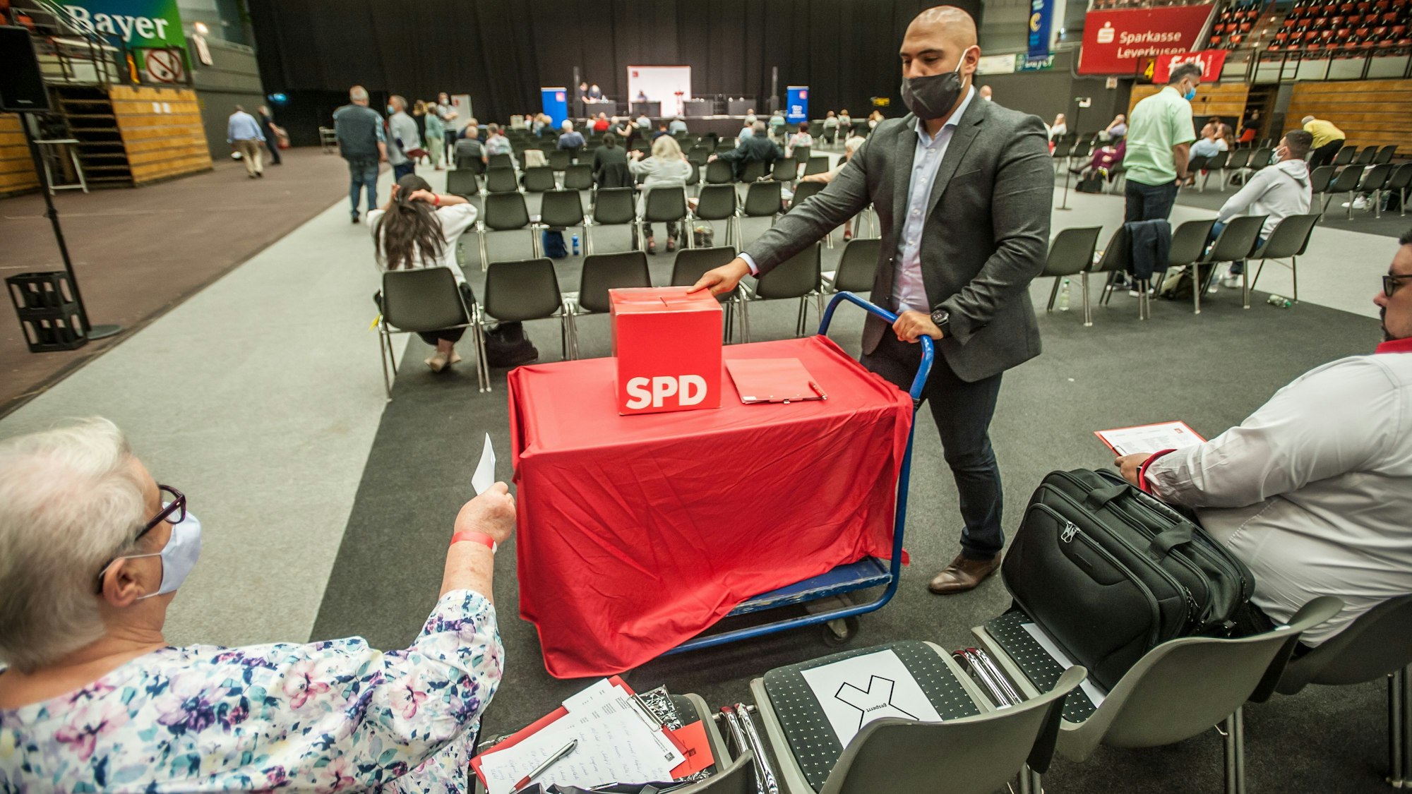 In einer großen Halle sitzen vereinzelt Menschen. Ein Mann schiebt einen kleinen Wagen durch die Sitzreihen. Darauf steht eine Wahlurne mit SPD-Logo.