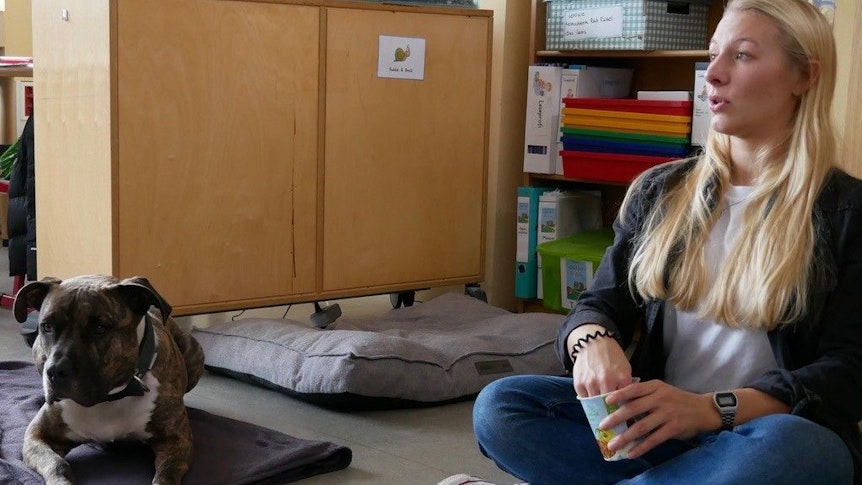 Die Fachlehrerin Vivienne Caspari sitzt neben ihrem Hund Merlin auf dem Boden des Klassenraums.
