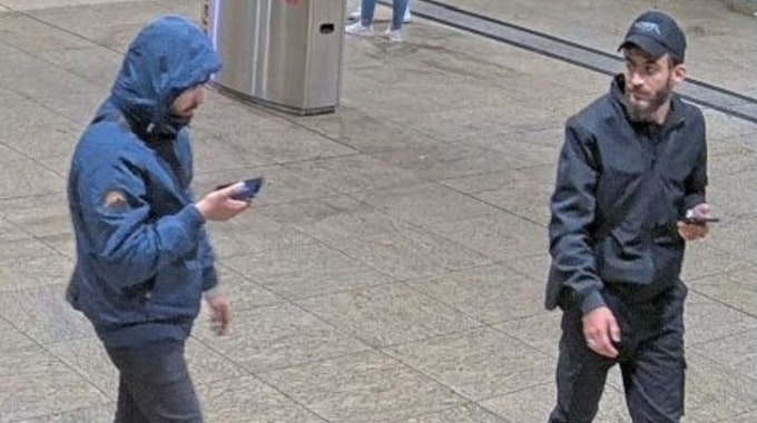 Zwei Männer gehen durch den Bahnhof. Beide halten ein Handy in der Hand. Einer trägt eine blaue Jacke mit aufgesetzter Kapuze. Der andere trägt eine schwarze Jacke samt schwazer Basecap.