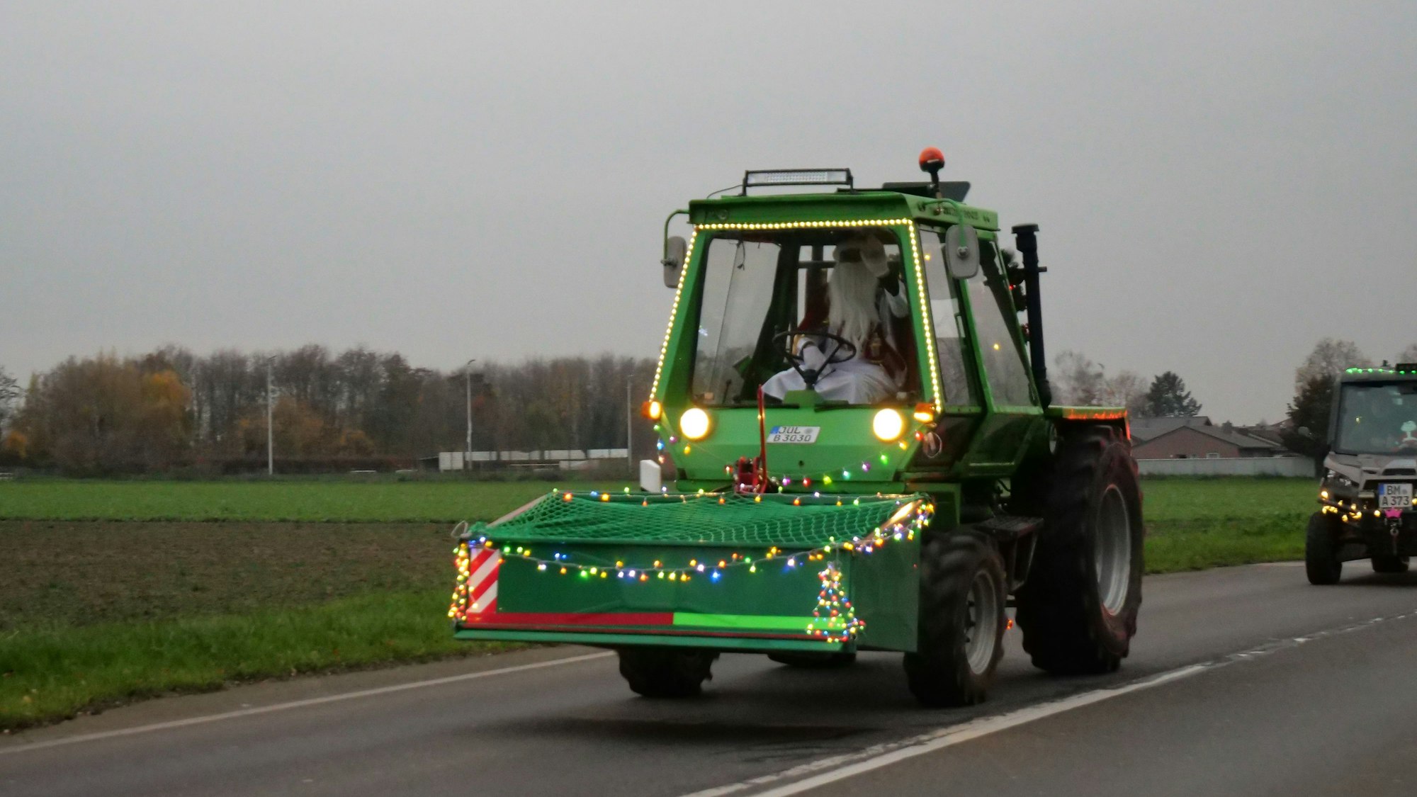 Ein geschmückter, grüner Traktor fährt über eine Straße. Am Steuer sitzt ein Mann, der als Weihnachtsmann verkleidet ist.