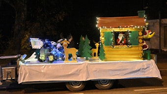 Auf einem Traktoranhänger ist ein Blockhaus mit Weihnachtsmann, Rentier und Traktor zu sehen.