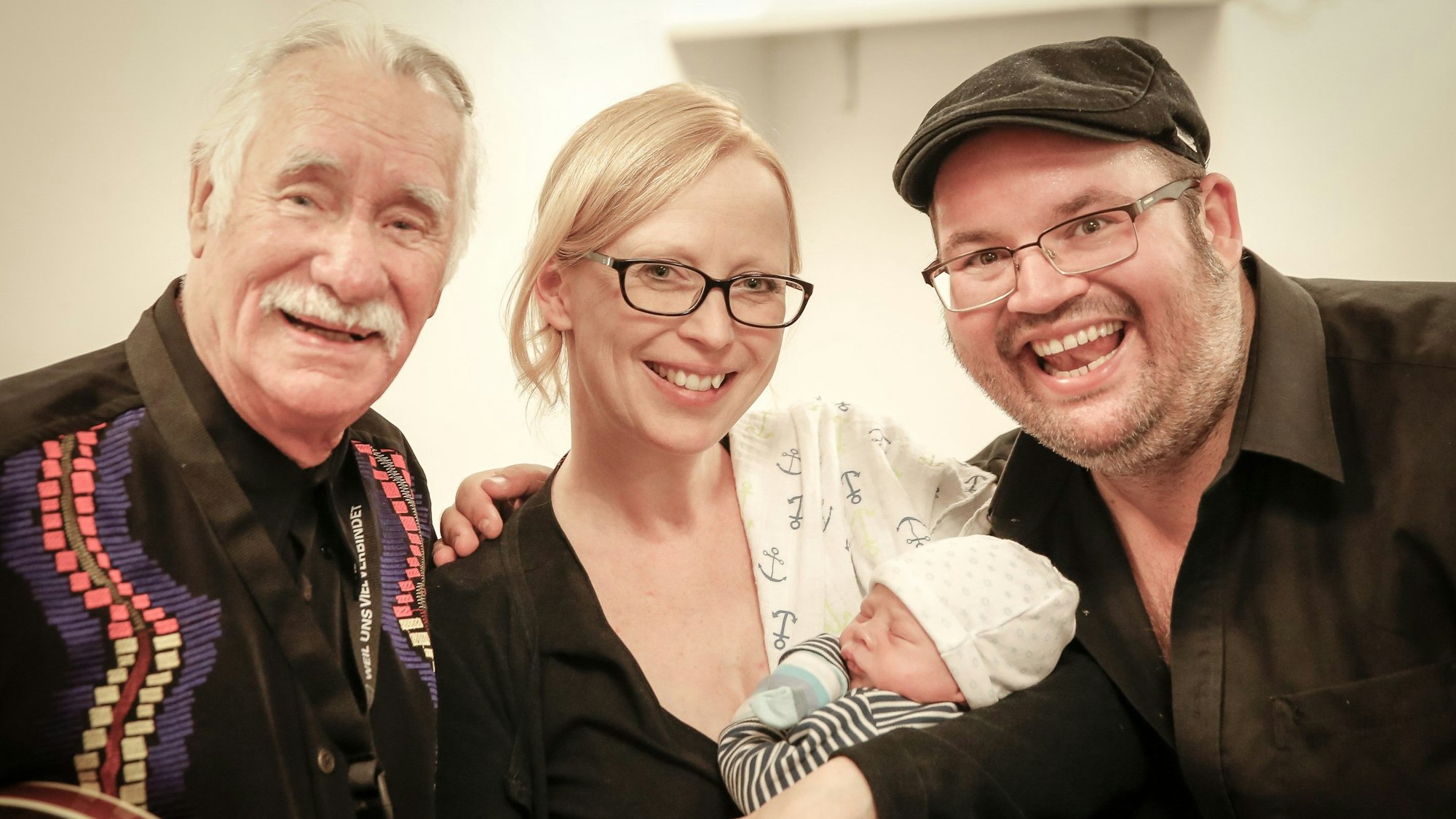 Hans Süper lächelt mit Björn Heuser und dessen Ehefrau Frau Iris, die ihr Baby im Arm hält, in die Kamera.