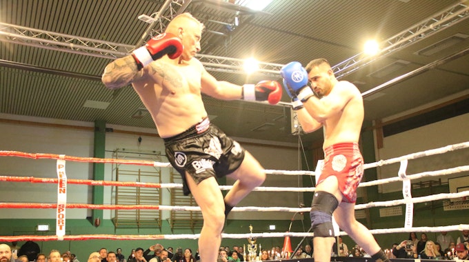 Zwei Kickboxer kämpfen gegeneinander im Ring, im Hintergrund befindet sich ein gut gefüllter Zuschauerraum.