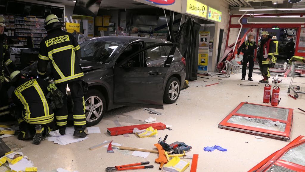 Ein Wagen ist in ein Einkaufszentrum in Dortmund-Aplerbeck gerast und kam dort zum Stehen. Feuerwehrkräfte kümmern sich um das Auto. Auf dem Boden liegen zerbrochene Gegenstände.